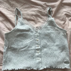 Ljusgrå ribbad linnetopp med spets - Supersöt ljusgrå ribbad linnetopp från Censored med spetskant vid v-ringningen, knappar framtill och små volangdetaljer längst ner. Axelbanden är smala och toppen har en liten rosett vid etiketten. Perfekt för en chill och söt look.