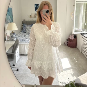 Vit studentklänning från NA-KD - Superfin vit klänning från NA-KD med lång ärm och vackra broderade blommor i spets. Klänningen har v-ringning med knyt i nacken och volang nedtill. Perfekt för sommarens tillfällen! Storlek 42. Endast använd en gång på studenten. Väldigt fint skick.