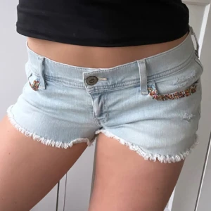 Ljusblå jeansshorts från Hollister - Säljer ett par ljusblå jeansshorts från Hollister med låg midja. Shortsen har fransiga benslut och färgglada detaljer vid fickorna framtill. Klassisk femficksmodell och slitningar på baksidan. Perfekta för varma dagar!