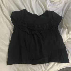 Svart blus med volang från Étoile Isabel Marant - Säljer en svart blus från Étoile Isabel Marant med volangdetaljer framtill. Blusen har rund halsringning och korta ärmar. Perfekt för dig som gillar stilrena plagg med en twist.