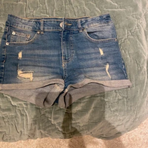 Blå jeansshorts med slitningar - Snygga blå jeansshorts med slitna detaljer och uppvikta benslut. Klassisk femficksmodell med knapp och dragkedja framtill. Perfekta för varma dagar och enkel att matcha med olika toppar.