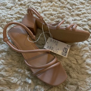 Beiga sandaler med klack från H&M - Säljer ett par stilrena beiga sandaler från H&M med fyrkantig tå och låg klack. Skorna har tunna remmar och öppen häl, perfekta för varmare dagar. Snygg och enkel design som passar till många outfits. Storlek 35, helt nya och oanvända!