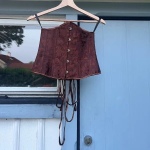 Brun korsett med snörning - Säljer en brun korsett med dekorativt mönster och snörning bak. Perfekt för att lyfta en outfit med en unik touch.