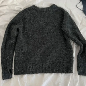 Mörkgrå stickad tröja - Mysig mörkgrå stickad tröja med rund halsringning och långa ärmar. Tröjan har en klassisk rak passform och ribbade muddar vid ärmslut och nederkant. Perfekt för kyliga dagar.