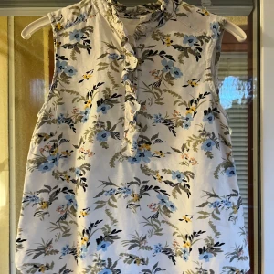 Blommig ärmlös blus med volangdetalj - Söt ärmlös blus med blommigt mönster i blått, gult och grönt på vit botten. Volangkant längs knäppningen framtill och liten volang vid halsen.