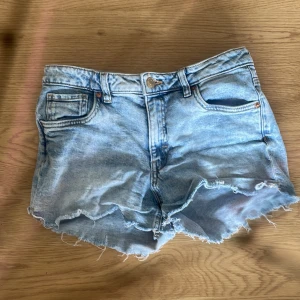 Ljusblå jeansshorts med fransar - Säljer ett par ljusblå jeansshorts från C&A med råa franskantade benslut och klassisk femficksmodell. Perfekta för sommardagar och har en avslappnad vibe. Stängs med knapp och dragkedja framtill.