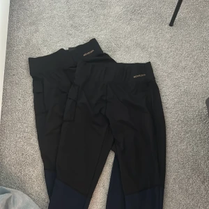 Svarta leggings - Säljer 2 par svarta leggings med en praktisk ficka på sidan. Byxorna har en tight passform och är perfekta för träning eller vardagsbruk. Materialet känns stretchigt och bekvämt.