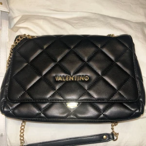 Svart quiltad axelväska från Valentino - Säljer en svart quiltad axelväska från Valentino med guldfärgad kedja och logga framtill. Väskan har en klassisk design och är perfekt för att lyfta vilken outfit som helst.