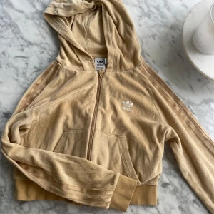 Beige velour hoodie från Adidas - Säljer en beige hoodie i velour från Adidas med dragkedja och huva. Jackan har de klassiska tre ränderna längs ärmarna och broderad logga på bröstet. Perfekt för en avslappnad och sportig stil. 