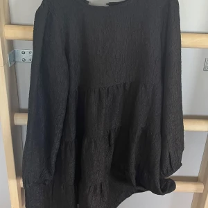 Svart långärmad klänning från Lindex - Svart klänning från Lindex med lång ärm och lätt strukturerat tyg. Modellen är lös och har en avslappnad passform med volangdetaljer. Perfekt för dig som gillar enkel men stilren design.