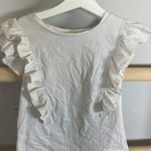 Vit topp med volangdetaljer från MNG - Säljer en söt vit topp från MNG med volangprydda axlar i spets. Toppen är ärmlös och har en rund halsringning. Perfekt för dig som gillar romantiska detaljer och en feminin stil.