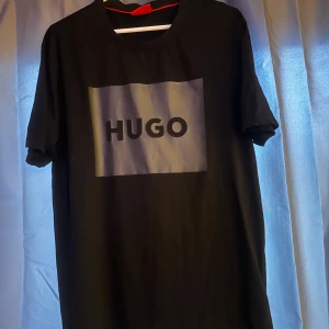 Svart t-shirt från Hugo - Säljer en svart t-shirt från Hugo med stor logga i grått och svart framtill. Klassisk rund hals och korta ärmar. Perfekt för en stilren och enkel look.