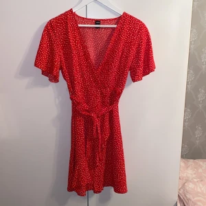 Röd omlottklänning med knyt från SHEIN - Superfin röd omlottklänning från SHEIN med vitt prickigt mönster. Klänningen har v-ringning, korta ärmar och knytband i midjan. Perfekt för sommarens alla tillfällen! Säljer då jag inte använder den och v-ringningen är lite för stor💕