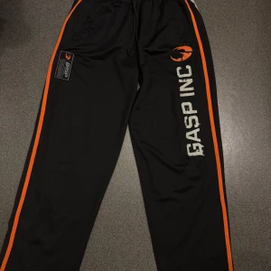 Svarta träningsbyxor från GASP INC   - Säljer ett par svarta träningsbyxor från GASP INC med orangea ränder längs sidorna och tryckt logga på ena benet. Byxorna har dragskedjefickor och raka ben, perfekta för gymmet.