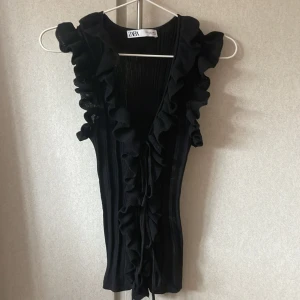 Svart volangtopp från Zara - Säljer en svart ärmlös topp från Zara med djup v-ringning och volangdetaljer längs kanterna. Toppen är ribbad och har en feminin, elegant look som passar perfekt till många olika outfits.