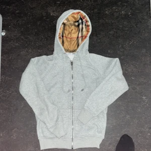 Grå hoodie från Burberry med klassiskt rutigt foder - Säljer en grå hoodie från Burberry med dragkedja och justerbar huva. Insidan av huvan har det ikoniska Burberry-rutiga fodret i beige, svart och rött. Perfekt för en stilren och avslappnad look. Passar xs/s