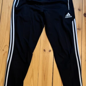 Svarta träningsbyxor från Adidas med vita ränder - Svarta träningsbyxor från Adidas med klassiska vita ränder längs sidorna och Adidas-logga på låret. Byxorna har dragkedjor vid fickorna och är tillverkade i ett lätt och svalt material med climacool-teknologi. Storlek M.