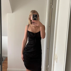 Svart satin klänning med smala axelband - Säljer en svart klänning i satin med smala axelband och knytdetalj framtill. Klänningen har en figurnära passform och är kort i modellen, perfekt för dig som gillar en stilren och enkel look.