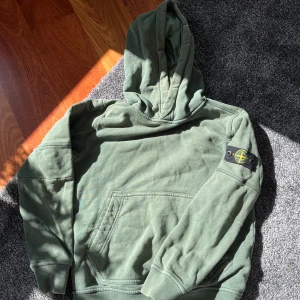 Grön hoodie från Stone Island Junior storlek 10y - Säljer en grön hoodie från Stone Island Junior med klassisk patch på ärmen. Tröjan har huva, magficka och ribbade muddar. Perfekt för en avslappnad och cool stil.