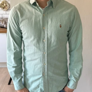 Ljusgrön skjorta från Ralph Lauren - Säljer en ljusgrön skjorta från Ralph Lauren med klassisk button-down krage och broderad logga på bröstet. Skjortan har långa ärmar och knäppning framtill. Perfekt för en stilren look! Dm för frågor🙌
