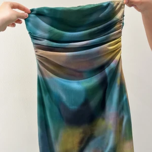 Grön axelbandslös långklänning - Säljer en snygg axelbandslös långklänning i gröna och blå toner med draperad överdel. Klänningen har ett mjukt, skimrande material och ett unikt mönster som ger en cool look. Perfekt för dig som vill sticka ut!