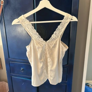 Vit satinlinnetopp med spetsdetaljer - Säljer en elegant vit linnetopp i satin med breda spetsband och vacker spets längs hela framsidan. Toppen har en djup v-ringning och är ärmlös, perfekt för en stilren look.