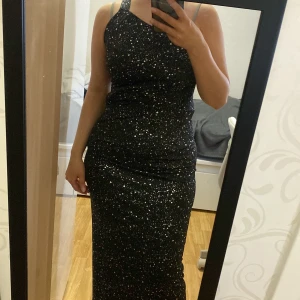 Svart glittrig långklänning med one shoulder - Säljer en svart långklänning med glittriga detaljer och one shoulder-design. Klänningen har en tight passform och är perfekt för festliga tillfällen. Materialet är stretchigt och sitter snyggt längs kroppen.