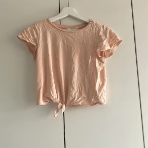 Ljusrosa topp med spetsdetaljer från H&M - Söt ljusrosa topp från H&M med korta ärmar i spets och knytning framtill. Toppen har rund halsringning och är tillverkad i mjuk bomull. Perfekt för en somrig look med fina detaljer på ärmarna.