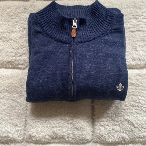 Marinblå stickad tröja med dragkedja från Morris - Säljer en marinblå stickad tröja från Morris med halv dragkedja och ribbad krage. Tröjan har en liten broderad logga på bröstet och är perfekt för lager-på-lager. Klassisk och stilren design. Nypris 2000kr