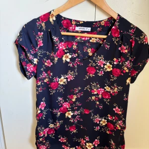 Mörkblå blommig blus från MyHailys - Säljer en mörkblå blus från MyHailys med färgglatt blommönster i rött, gult och vitt. Blusen har korta ärmar och en v-ringad halsringning. Perfekt för dig som gillar färg och mönster!