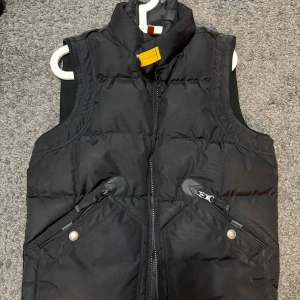 Svart dunväst från Parajumpers - Säljer en svart dunväst från Parajumpers med hög krage och gul detalj vid halsen. Västen har två snedställda fickor med dragkedja framtill och är vadderad för extra värme. 