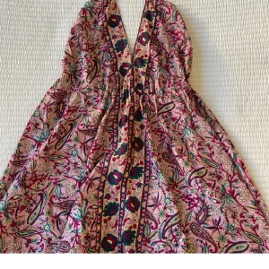 Blommig omlottklänning med knyt - Säljer en färgglad omlottklänning med blommigt mönster i rosa, grönt och beige. Klänningen har smala axelband, v-ringning och knyt i midjan. Perfekt för varma dagar och sommarens alla tillfällen.