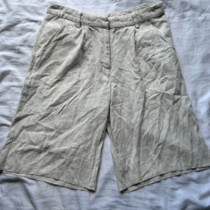 Beige linneshorts med vida ben - Säljer ett par beige shorts i linne med vida ben och hög midja. De har hällor för bälte, fickor fram och en dekorativ bakficka. Perfekta för varma dagar och ger en avslappnad men stilren look.