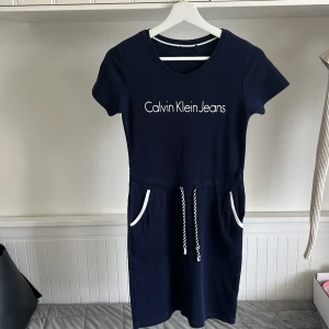 Marinblå klänning från Calvin Klein Jeans - Snygg marinblå klänning från Calvin Klein Jeans med vit logga framtill. Klänningen har korta ärmar, dragsko i midjan och vita detaljer vid fickorna. Perfekt för en avslappnad och sportig stil.