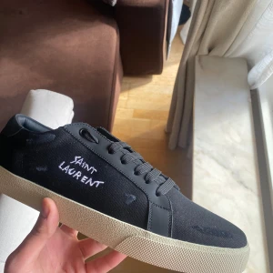 Svarta sneakers från Saint Laurent - Säljer ett par svarta sneakers från Saint Laurent med vit broderad logga på sidan. Skorna har snörning, rund tå och en beige platt sula. Ovandelen är i textil och detaljer i skinn. Perfekta för en stilren look. OBS Intressekoll