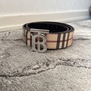 Beige och svart rutigt bälte från Burberry - Säljer ett klassiskt bälte från Burberry med beige och svart rutigt mönster samt silverfärgat BB-spänne. Bältet har inslag av rött och vitt i mönstret och är tillverkat i skinn.