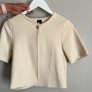 Beige kortärmad cardigan jacka från Gina Tricot - Säljer en stilren beige cardigan jacka från Gina Tricot. Jackan har korta ärmar, rund halsringning och stängs med en dekorativ knapp framtill. Perfekt för lager-på-lager och enkel att matcha.