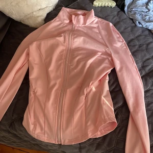 Ljusrosa träningsjacka med dragkedja - Säljer en ljusrosa träningsjacka med hel dragkedja framtill och hög krage. Jackan har långa ärmar och två fickor med dragkedja på sidorna. Perfekt för dig som vill ha en sportig och stilren look.