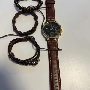 Klocka med brunt läderarmband och guldboett - Stilren klocka med mörkblå urtavla, romerska siffror i guld och tre mindre urtavlor. Boetten är guldfärgad och armbandet är i brunt skinn med vita sömmar. Perfekt accessoar för dig som gillar klassisk design.