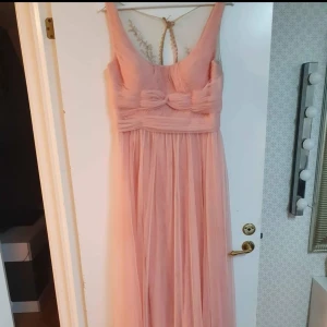 Lång rosa klänning med spets och rosett - Romantisk långklänning i ljusrosa med vacker spets upptill och dekorativa knappar i ryggen. Klänningen har en söt rosettdetalj framtill och transparent mesh med broderade blommor på ryggen. Perfekt för bal eller fest.