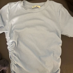 Ljusblå ribbad topp-zara - Säljer en ljusblå ribbad topp från Zara med korta ärmar och rund halsringning. Toppen har en snygg rynkdetalj med snörning på sidorna. 