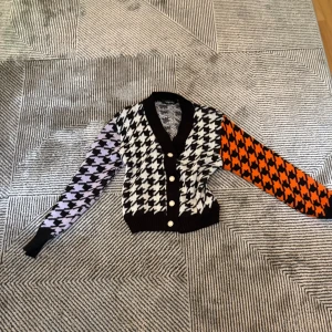 Mönstrad kofta från NAANAA - Färgstark kofta från NAANAA med stora hundtandsmönster i svartvitt, lila och orange. Koftan har svarta ribbade muddar, v-ringning och knappar framtill. Perfekt statement-plagg för dig som gillar unika detaljer.