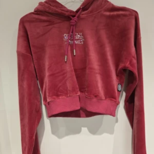 Rosa croppad velour hoodie från Von Dutch - Säljer en rosa croppad hoodie och vida byxor från Von Dutch i mjuk velour med glittrande logga framtill. Tröjan har huva med dragsko och långa ärmar. Perfekt för dig som gillar en trendig och bekväm stil.