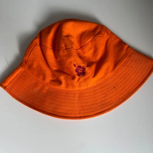 Orange bucket hat med blommönster - Cool orange bucket hat med lila broderad blomma framtill. Insidan har ett färgglatt blommigt mönster i orange och lila. Köpte för runt 100kr (?) på ale-hop