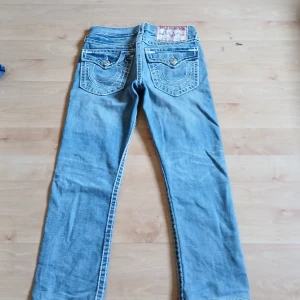 True Religion ljusblå jeans med kontrastsömmar - Säljer ett par ikoniska ljusblå jeans från True Religion med tydliga vita kontrastsömmar och stora fickor bak med lock och knapp. Klassisk femficksmodell och läderpatch bak i midjan. Perfekta för dig som gillar streetwear och vill sticka ut.