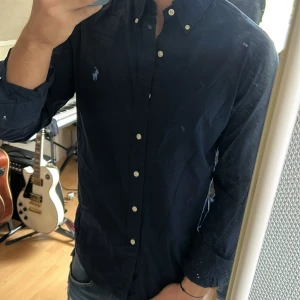 Mörkblå skjorta från Ralph Lauren - Säljer en klassisk mörkblå skjorta från Ralph Lauren med vit broderad logga på bröstet. Skjortan har knappar hela vägen, button-down krage och långa ärmar. Perfekt till jeans eller chinos för en clean look.