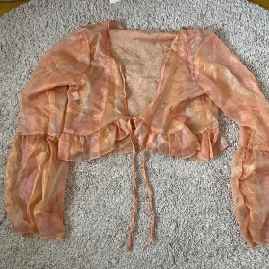 Glansig rosa bolerotopp med volang och knyt - Säljer en transparent och glansig orange bolerotopp med långa ärmar, volangkant och knyt framtill. Perfekt för att lyfta en outfit med sitt skimrande material och lätta känsla. Använd endast en gång. Lite ABBA-Vibe. Gissar storlek M
