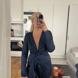 Marinblå omlottklänning med v-ringning - Säljer en marinblå omlottklänning från Missguided med lång ärm och djup v-ringning. Klänningen har en snygg knutdetalj framtill och är tillverkad i ett glansigt, satinliknande material. Perfekt för dig som vill ha en stilren och trendig look.