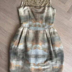 Champagnefärgad klänning med pärldetaljer - Säljer en elegant kortklänning i champagnefärg med skimrande pärlor upptill. Klänningen har ett unikt mönster i beige, grått och guldtoner samt en figurnära passform med veckad kjol. Perfekt för speciella tillfällen.