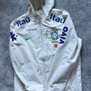 Beige Brasilien fotbollsjacka från Nike - Säljer en vit Nike jacka med Brasilien CBF-logga och sponsormärken. Jackan har huva, dragkedja och tryck i blått, grönt och gult. Perfekt för fotbollsälskare som vill ha något unikt och sportigt.